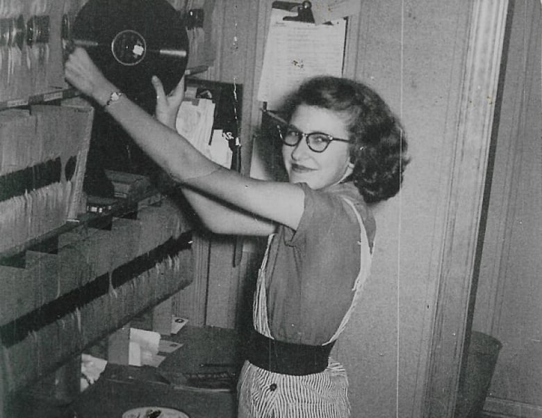 A black and white photo of a young, bespectacled woman smiling for the camera as she fetches a record from a shelf.