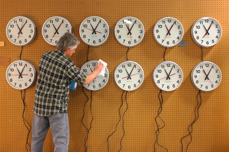 A man polishes the face of a clock hanging on a wall among 11 other clocks, all set to the same time. 