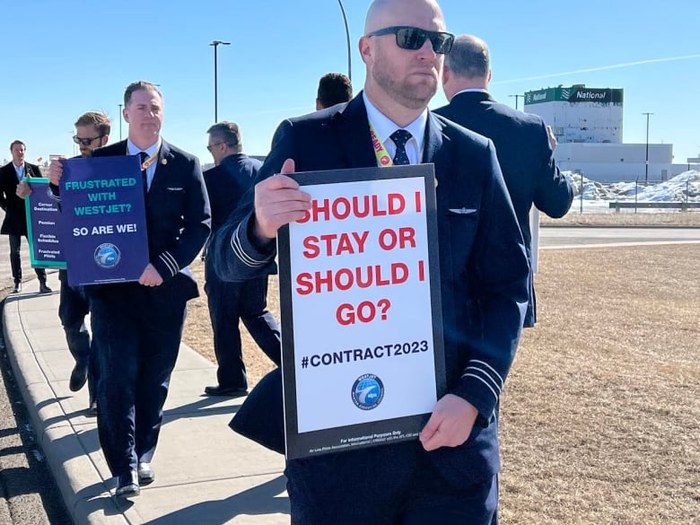 A bald man wearing sunglasses and a suit holds a sign that reads 'should I stay or should I go?.'