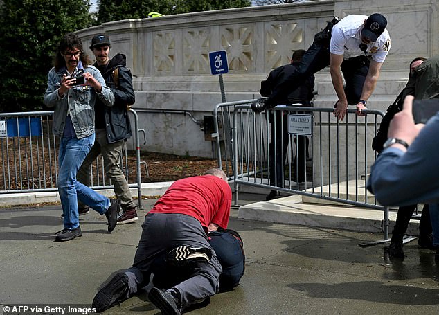 The man confronts one of the masked men holding the sign, who says that 'f***ots like you will try to get my job taken away.' Multiple people start to skirmish over the large sign before the man in the red shirt and the masked man begin to tussle