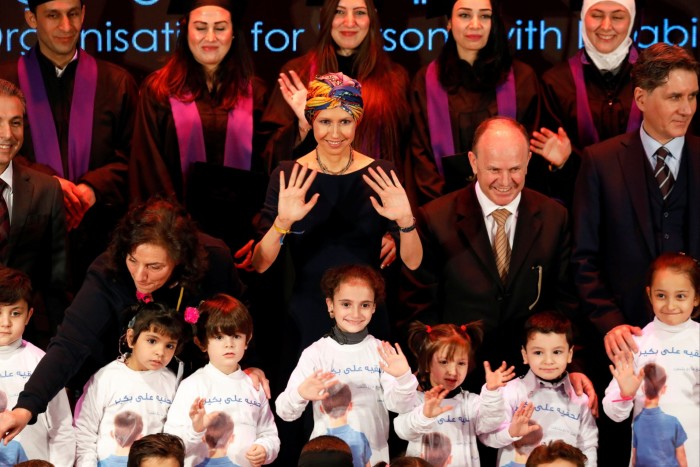 A woman in a multicoloured head covering stands amid a group of children and is flanked by university graduates in robes and mortar board hats