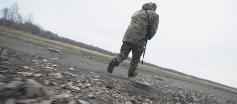 A man in military camouflage clothing and carrying a rifle runs away from the camera across a rural field dotted with landmines.