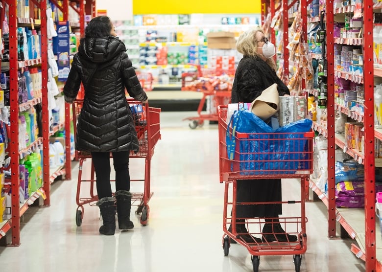 Two women with shopping carts walk through the aisle at a grocery store.