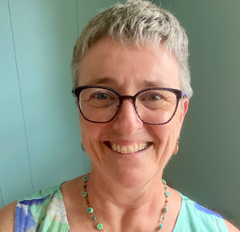 A woman with short hair and glasses smiles for a selfie in a room with aqua blue walls.