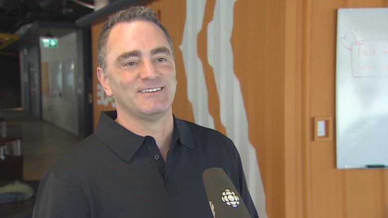 A white man wearing a black polo shirt speaks to a cameraman.