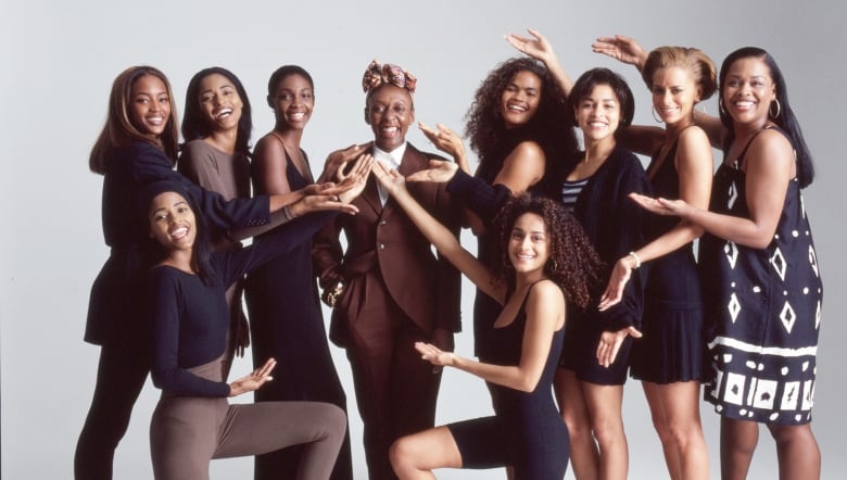 Nine smiling Black women gesture towards one older woman in the centre, who is grinning and sporting a chic brown suit.