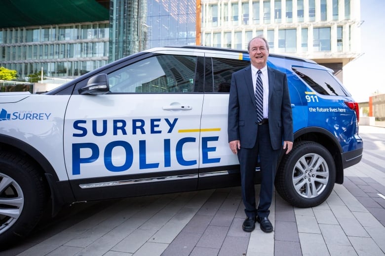 A smiling white man in a dark-blue suit stands beside an SUV emblazoned with the words "Surrey police."