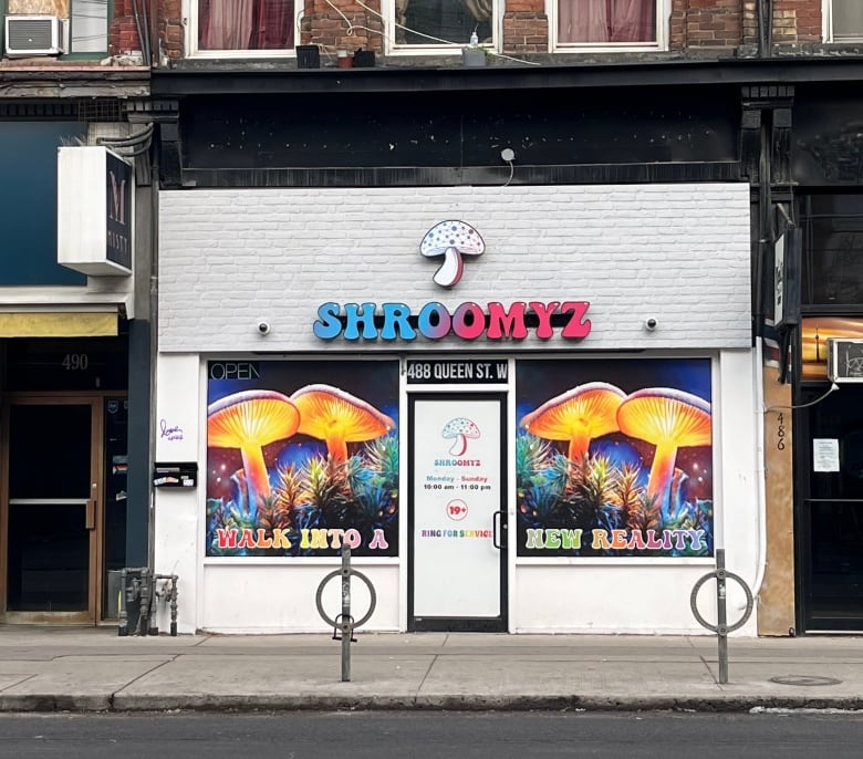 A mushroom logo and a sign that reads Shroomyz is pictured on a white brick storefront. 