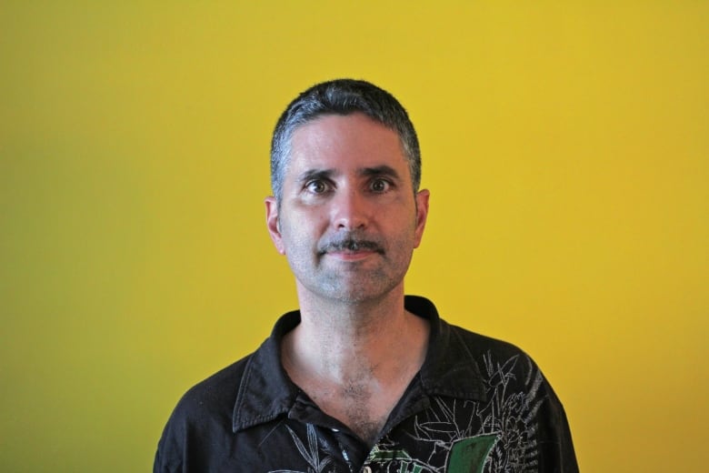 A man in a patterned black shirt poses for a photo against a bright yellow background.