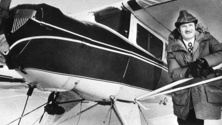 A man in a winter coat and fedora stands next to a small airplane.