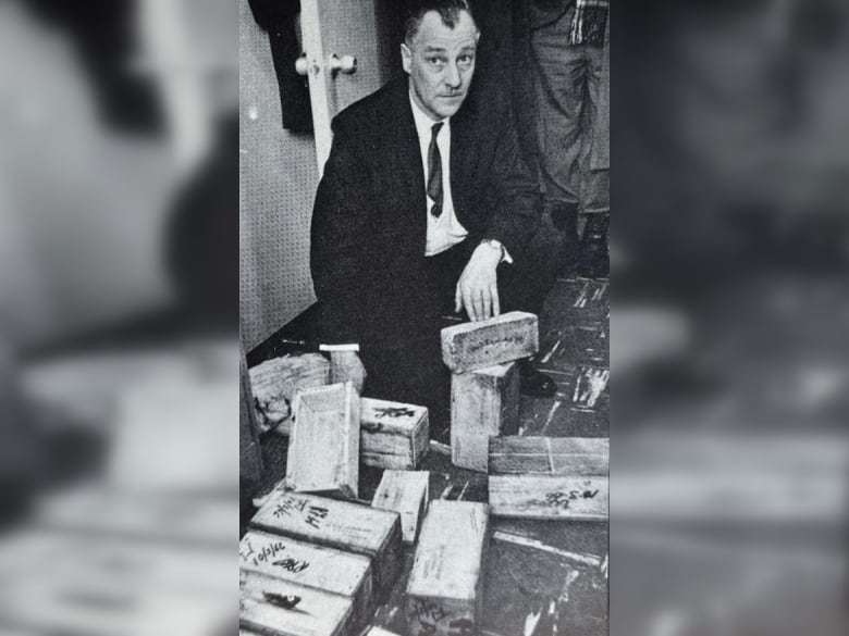 A balding man with grey hair and wearing a suit, squats down and poses with several rectangular bricks of gold.