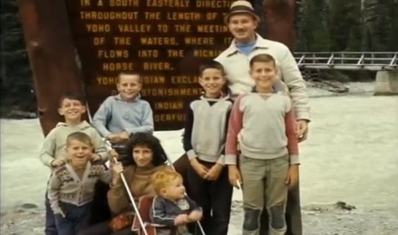 A family of eight, including a toddler in a push stroller, smile at the camera. The photo is coloured but slightly blurry.