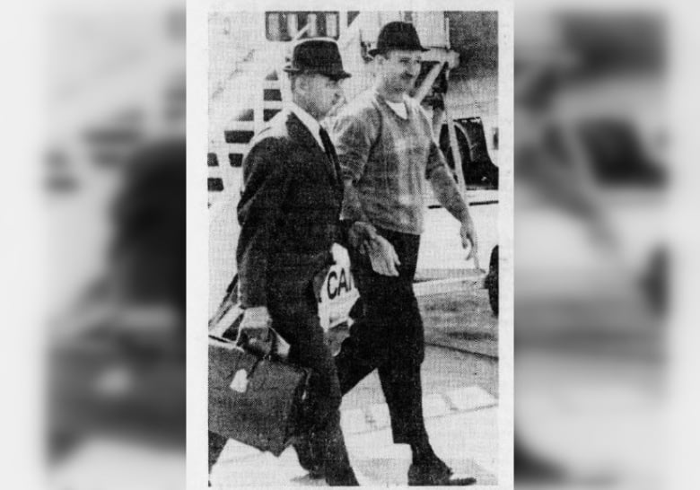 Two neatly dressed men walk side-by-side in a black and white photo from a newspaper. Both wear fedoras. One is in a suit and the other is in a sweater and slacks.