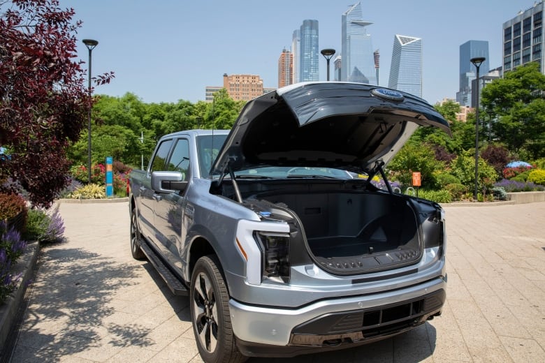 A silver truck with its front hood open, revealing an empty compartment, is parked on a driveway. In the background, a city skyline.