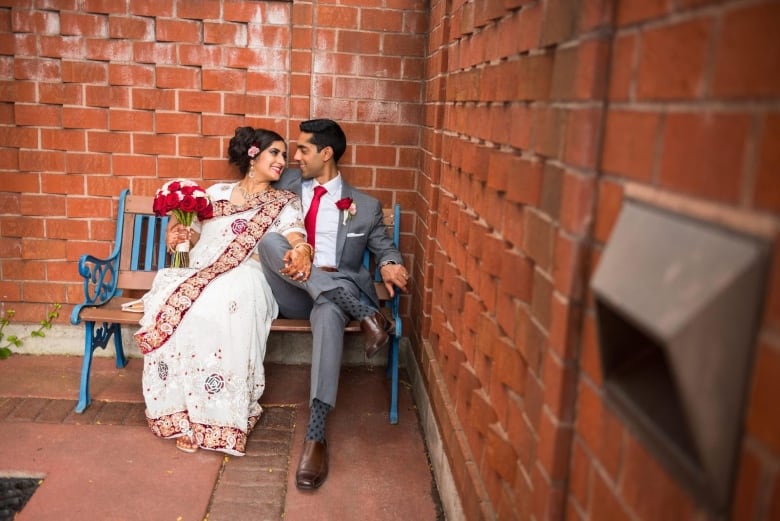 A woman in a white and red sari sits closely to a man in a suit as they lovingly look at each other. 