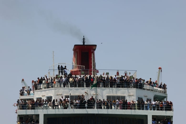 Smoke rises from a stack atop a large multi-storey ship packed with people, under a pale blue sky.