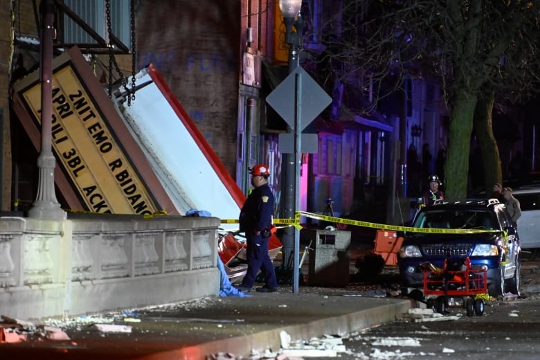 A marquis droops at an angle, with one end touching the ground near a city street. A man in a red helmet stands nearby.  The street and sidewalk are dirty.