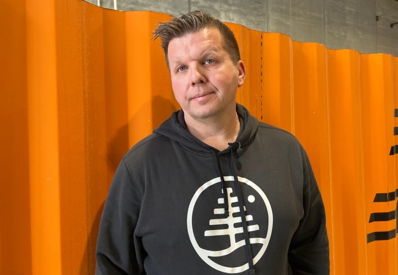 A man wearing a grey sweatshirt, with a white design on it, poses for a photo in front of an orange metal wall.