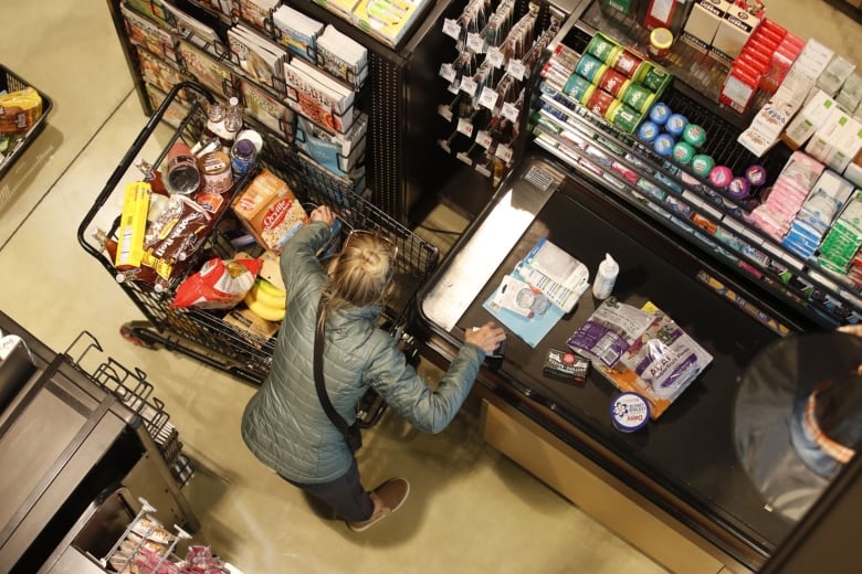 A person, seen from above, unloads items from a full grocery cart onto a conveyor belt at a cash register. 