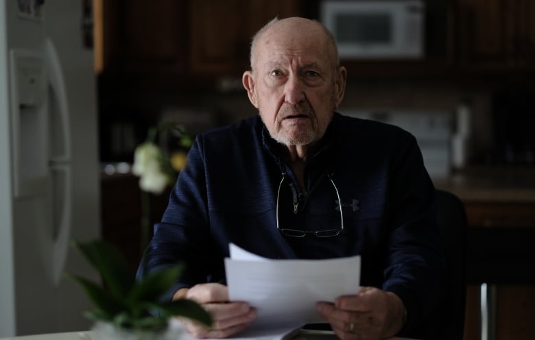 An older man with reading glasses around his neck sits at a table reading a handful of documents.