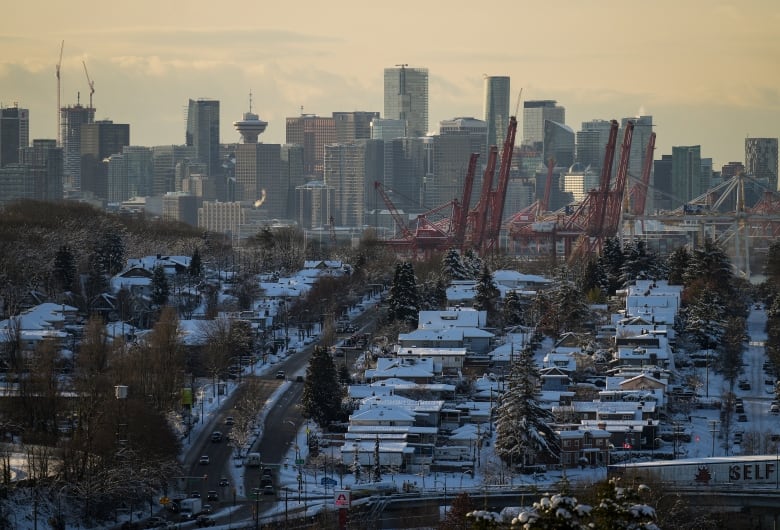 A shot of the Vancouver skyline looking North from South Vancouver. Image shows single family homes with snow-covered roofs in the foreground with the city skyline and North Shore mountains in the background. Many cranes are visible at various development sites.