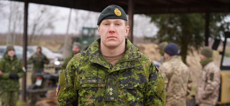 A young man in outdoor military camouflage clothing and a beret stands near a shelter where other soldiers are gathered.