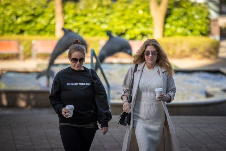 Two white women wearing sunglasses carrying coffee cups walk up a set of stairs towards an office building. A fountain with a dolphin is visible in the background.