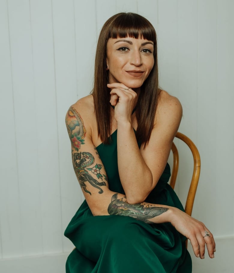 A woman with colourful tattoos on one arm wears a green one-shoulder gown and poses for a portrait on a chair. 