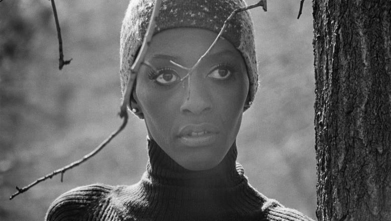 A black and white portrait of a Black woman in a hat peering from behind a tree branch.