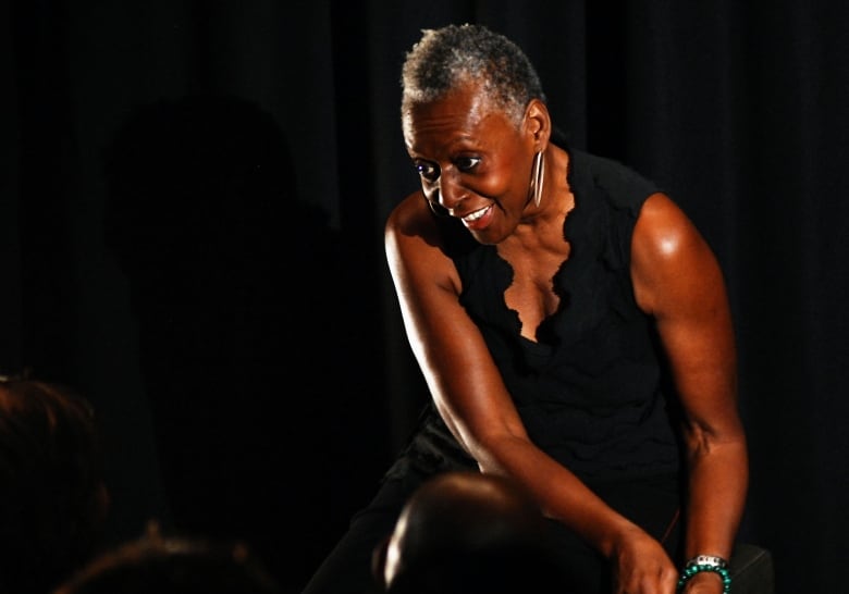 A woman in short grayish-white hair, seated on stage, smiles as she bends towards the audience. 