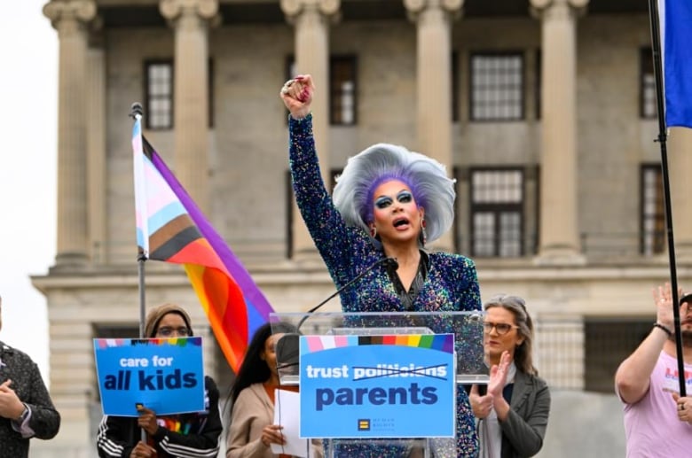 A drag queen raises her fist in the air at a podium.