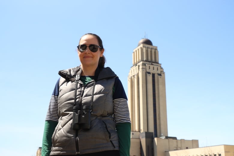 A woman with binoculars around her neck stands outside with a university tower in the background. 