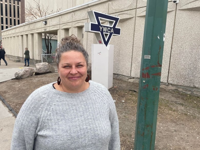 A smiling woman in a grey sweater stands outside a YWCA. 