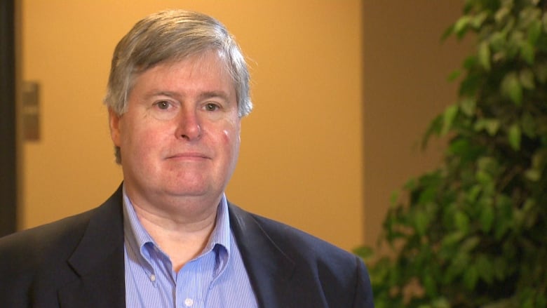 Ken Whitehurst, poses in front of a yellow wall with plants to his left.