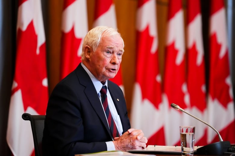 A man in a suit sits at some microphones in front of Canadian flags.