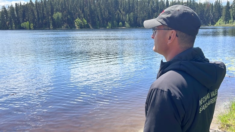 A man wearing a hoodie that says Nechako Valley Search and Rescue on the back looks over a tree-lined lake.