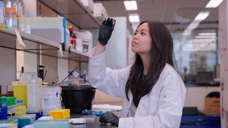 A woman in a white lab coat and black gloves inspects a small container.