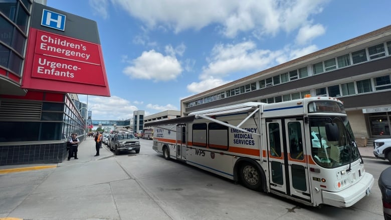 A large bus-like vehicle that says 'Emergency Medical Services' on the side is stopped on the street next to a building with a hospital sign that says 'Children's Emergency Urgence-Enfants.'