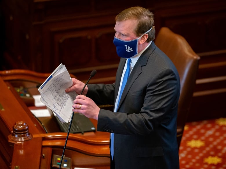 A man in a suit wearing a face mask stands in a legislature and speaks into a microphone while holding a stack of paper.