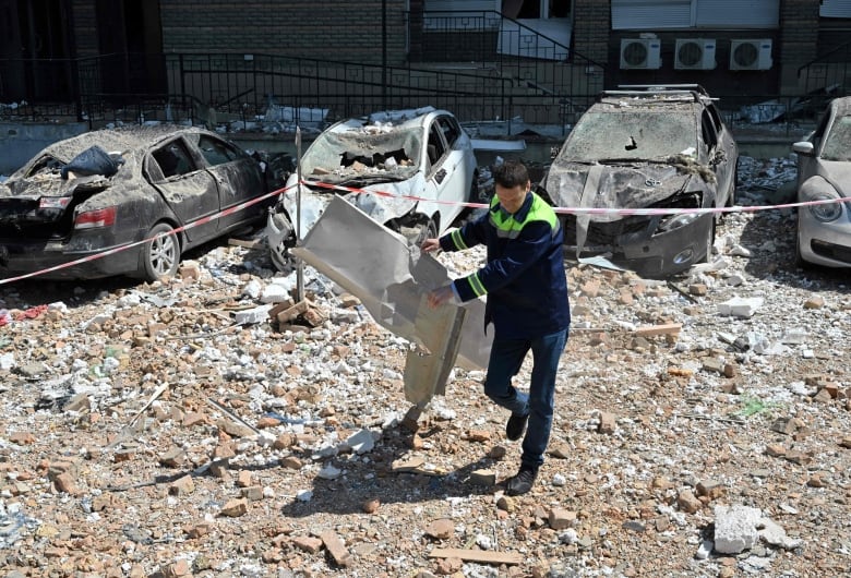 A man carries a large piece of debris as three heavily damaged automobiles are shown behind him.