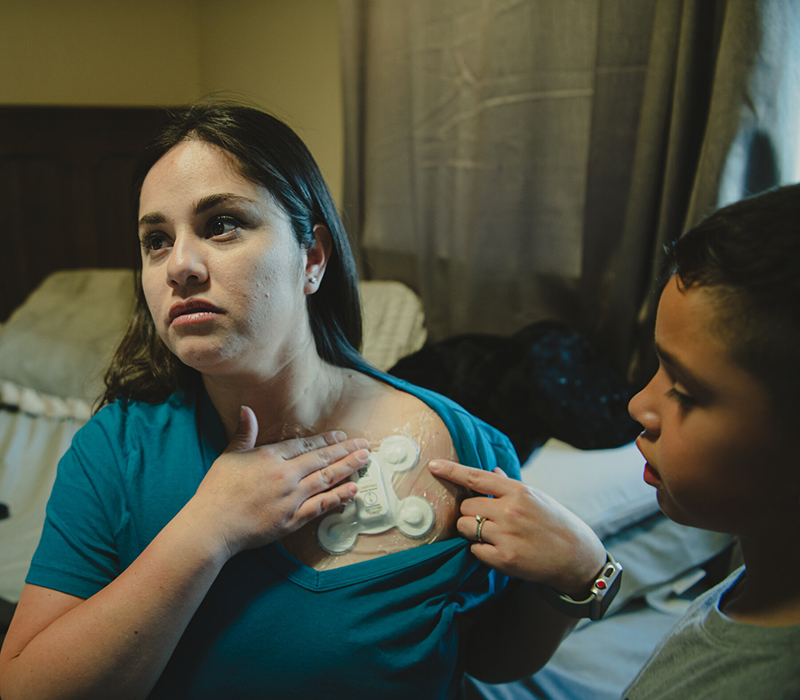 Ms. Rodríguez shows her young son the fist-sized biosensor she wears below her neck to monitor her heart. The sensor is white with four nodes branching off it.