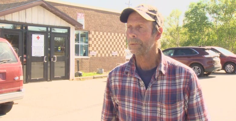 A man in a plaid shirt and ball camp stands in the parking lot in front of an evacuation centre.