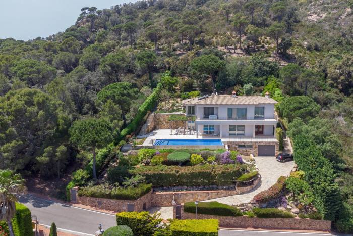 A two-storey home with paved curving driveway and pool on a sloping block with hill behind surrounded by scrubby forest