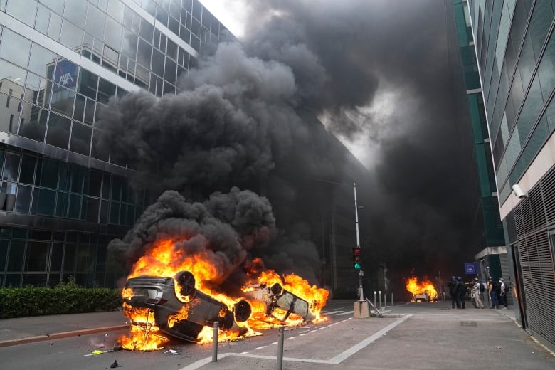 Black smoke arises from overturned, burning cars on the street.