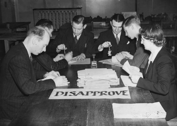 six men seated at a table sorting thorugh ballot papers. there is a sigh marked ‘DISAPPROVE’ on the table