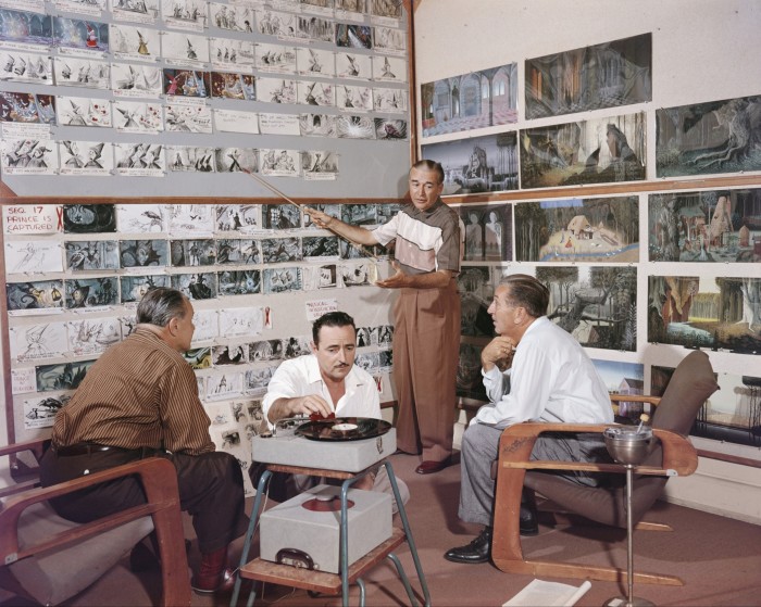 One man stands pointing to images pinned to a wall, while two men in chairs look on. Another man kneels on the floor, putting a record on a record player