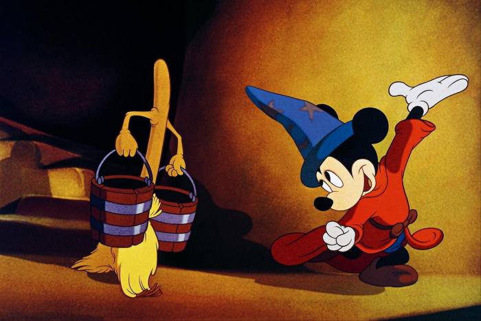 Mickey Mouse, in red coat and tall blue hat, stands in front of a broom that has been brought to life and is carrying buckets of water