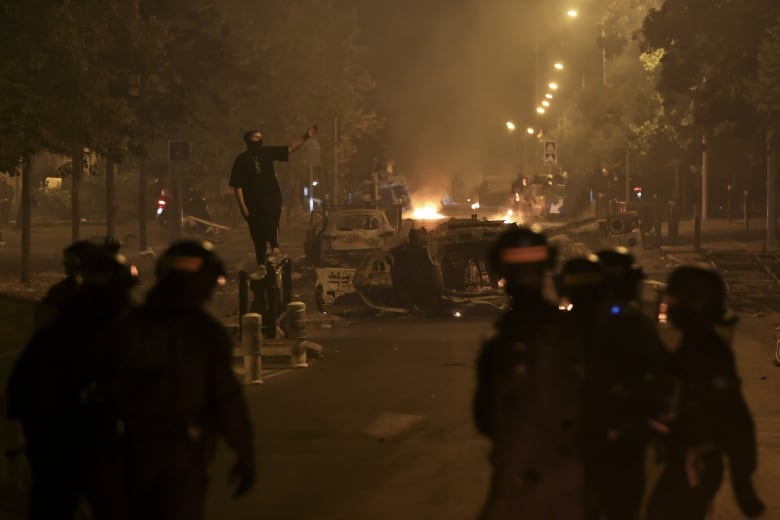 A fire burns in the distance on a street with several burned-out vehicles. A man with his face partially covered gestures as several people look on.