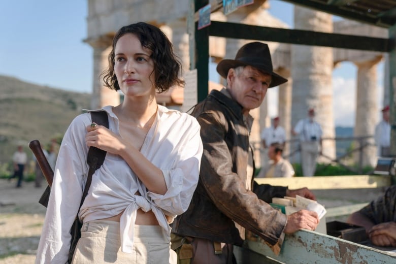 A woman stands to the left of a man. The woman is wearing a blouse tied in the middle. They are standing outside in front of what look to be ancient Greek ruins. 