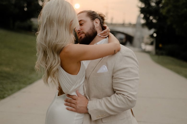 A man in a beige suit embraces a woman in a wedding dress in front of a bridge, beneath a hazy sky.